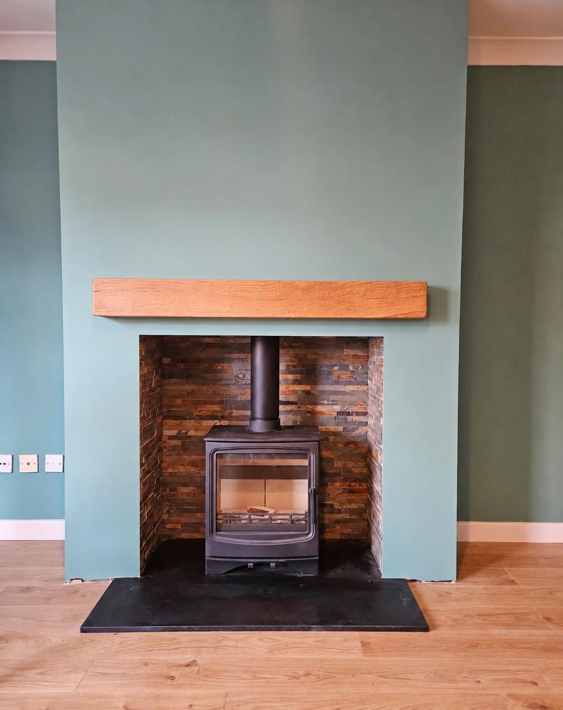 Black wood burner in a split-face stone inglenook with a floating oak beam and sage green walls